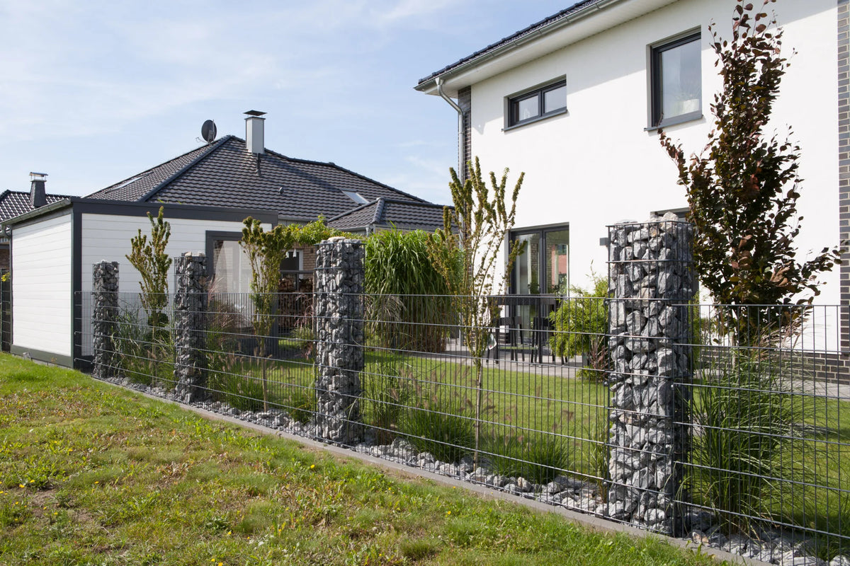Zaun mit Gabionen: Gabionensäulen RUBIN mit dunkler Steinfüllung zwischen Doppelstabmatten vor einem Einfamilienhaus